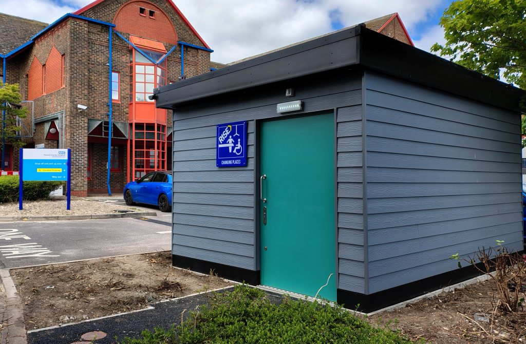 Changing Places facilitiy - outside Outdoor view of a grey utility building with a wheelchair-accessible symbol and an entrance beside a hospital with visible signage and parking spaces.
