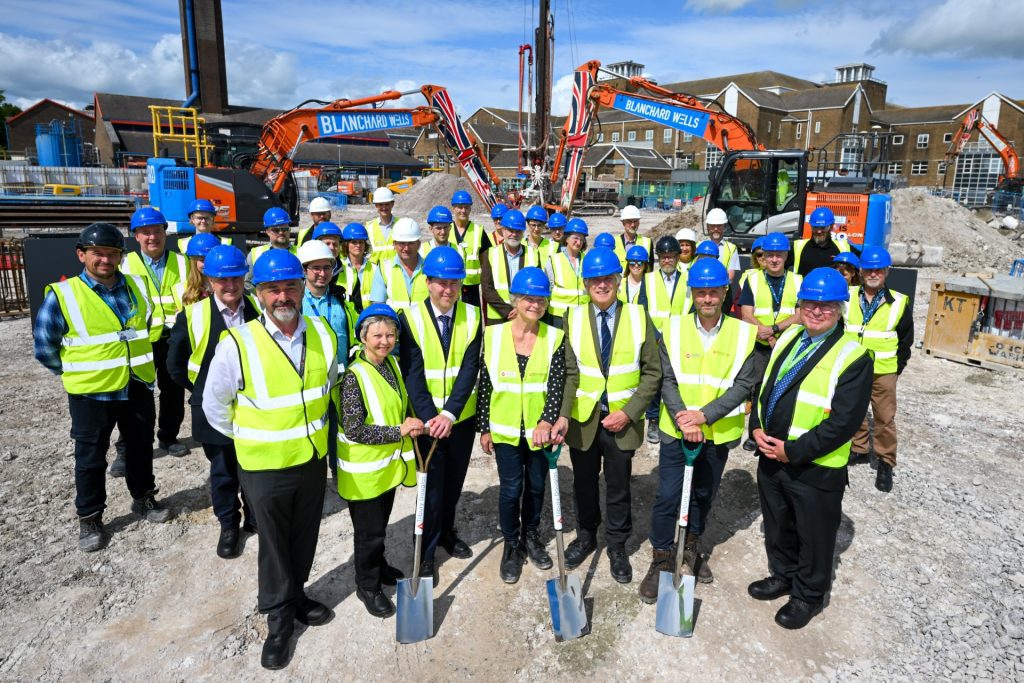 Groundbreaking group pic resize People in hard hats and high vis yellow jackets on a building site with spades in the ground and diggers in the background